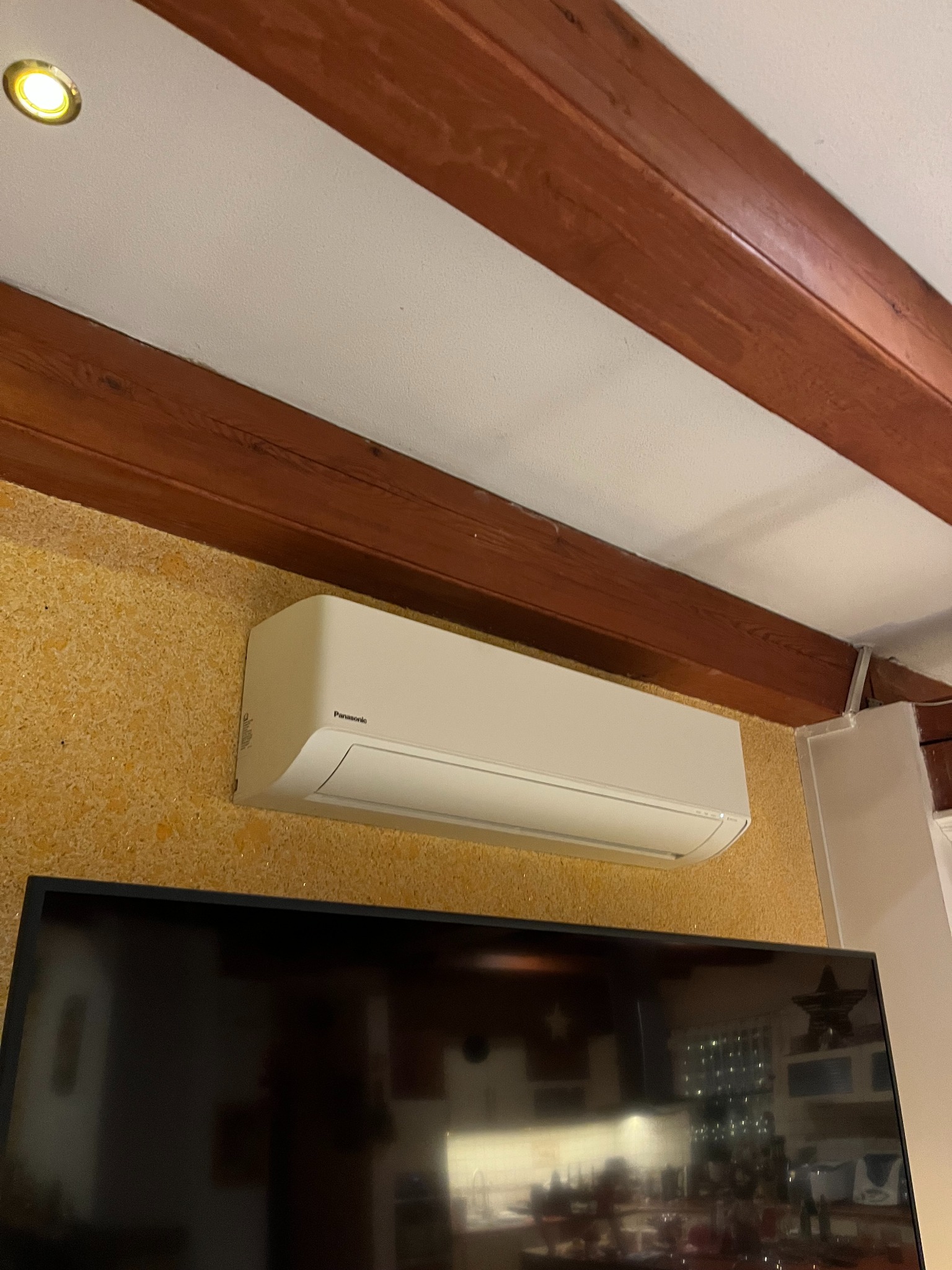 Klimatyzator Panasonic zamontowany na ścianie o fakturze korka, widoczny fragment telewizora i drewniane belki stropowe. Ujęcie z dołu.