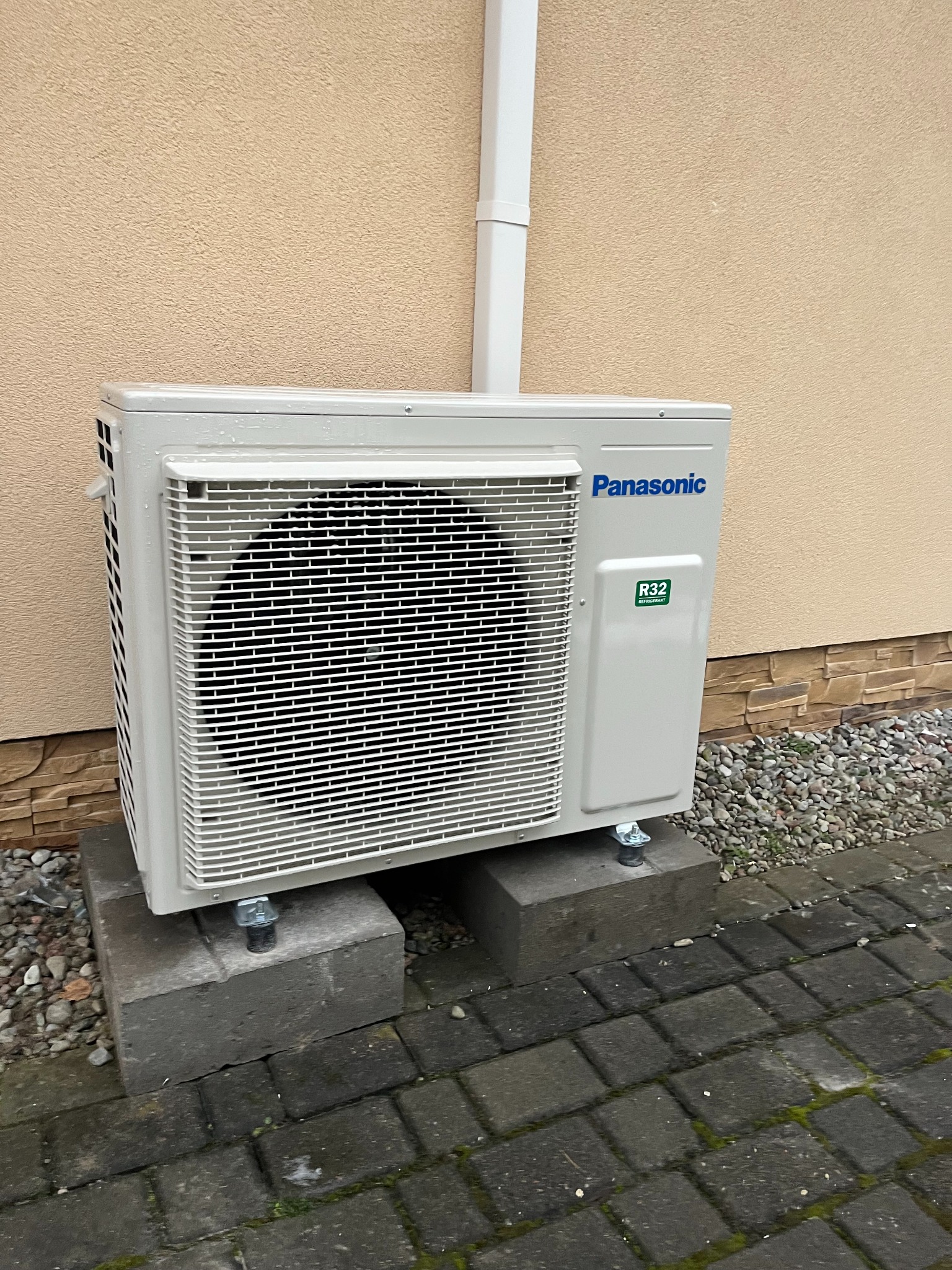 Zewnętrzna jednostka klimatyzacji Panasonic R32, zamontowana na betonowych blokach przy ścianie budynku z rurą spustową. Widoczne kostki brukowe i kamyczki.