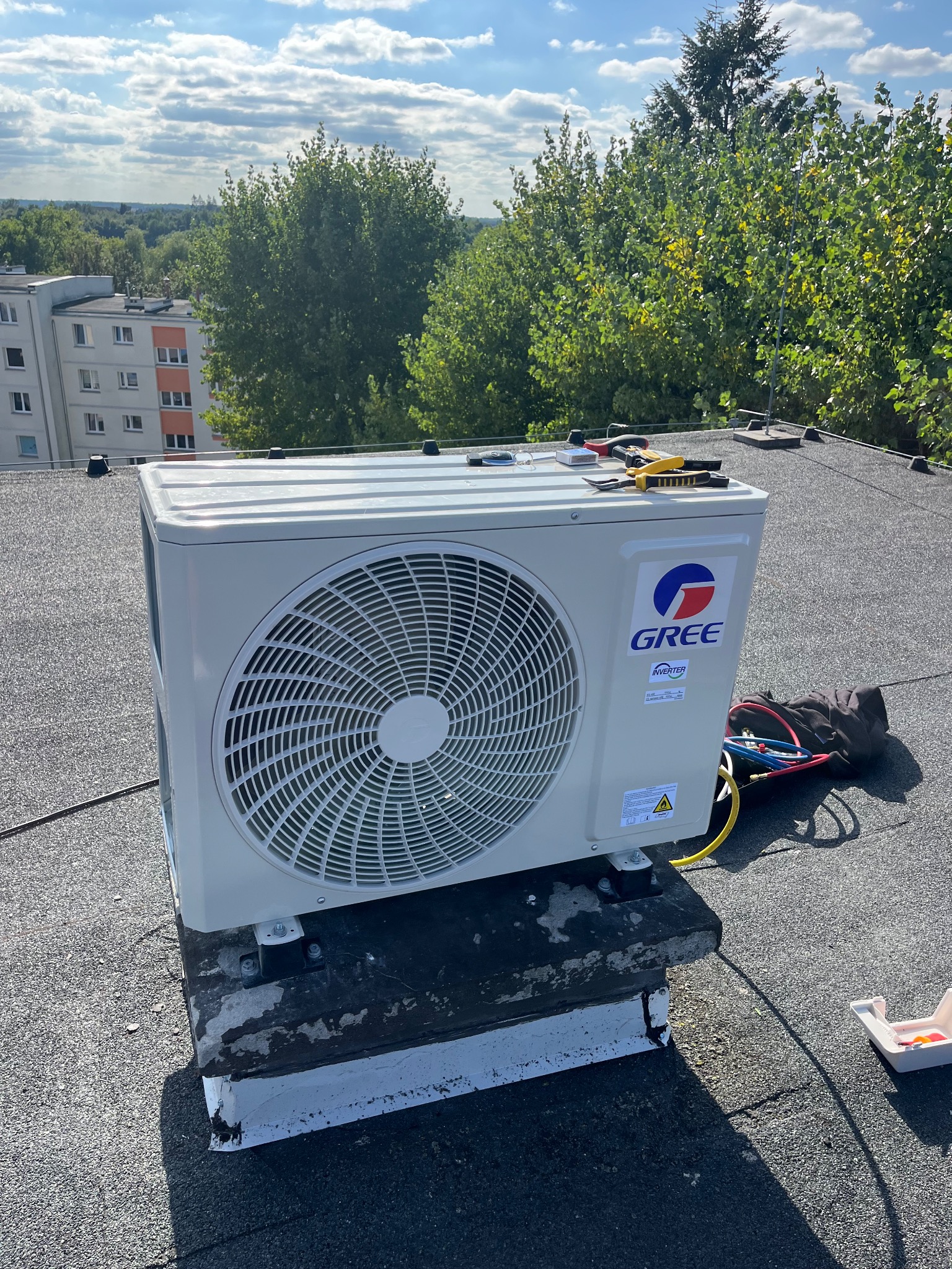 Biała jednostka zewnętrzna klimatyzacji marki Gree na dachu budynku, z narzędziami obok. Widok na zieleń i błękitne niebo w tle. Słoneczny dzień.