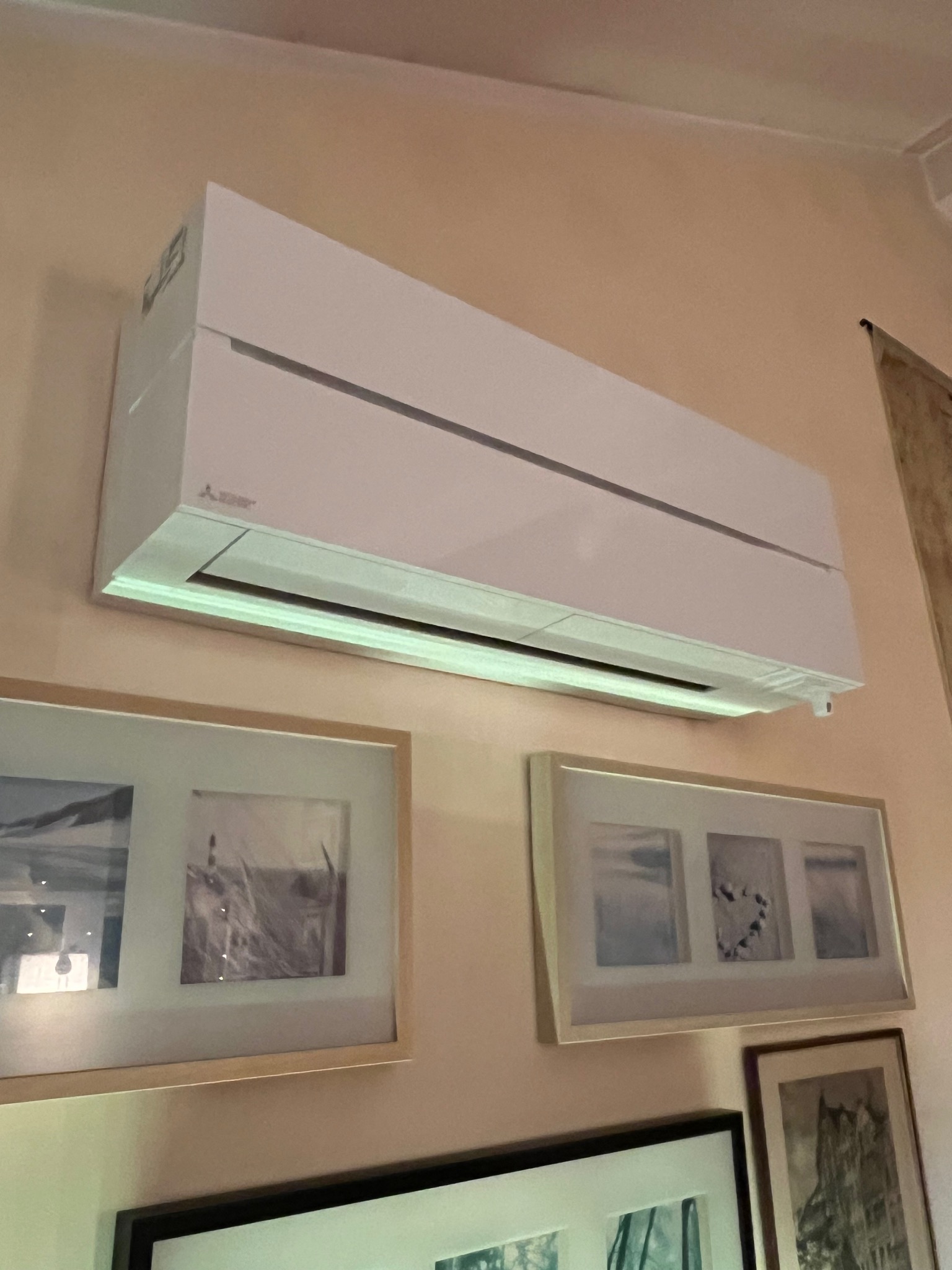 Biały klimatyzator Mitsubishi zamontowany na ścianie w pomieszczeniu, pod nim wiszą obrazy w ramkach. Widok z dołu, oświetlenie dzienne.