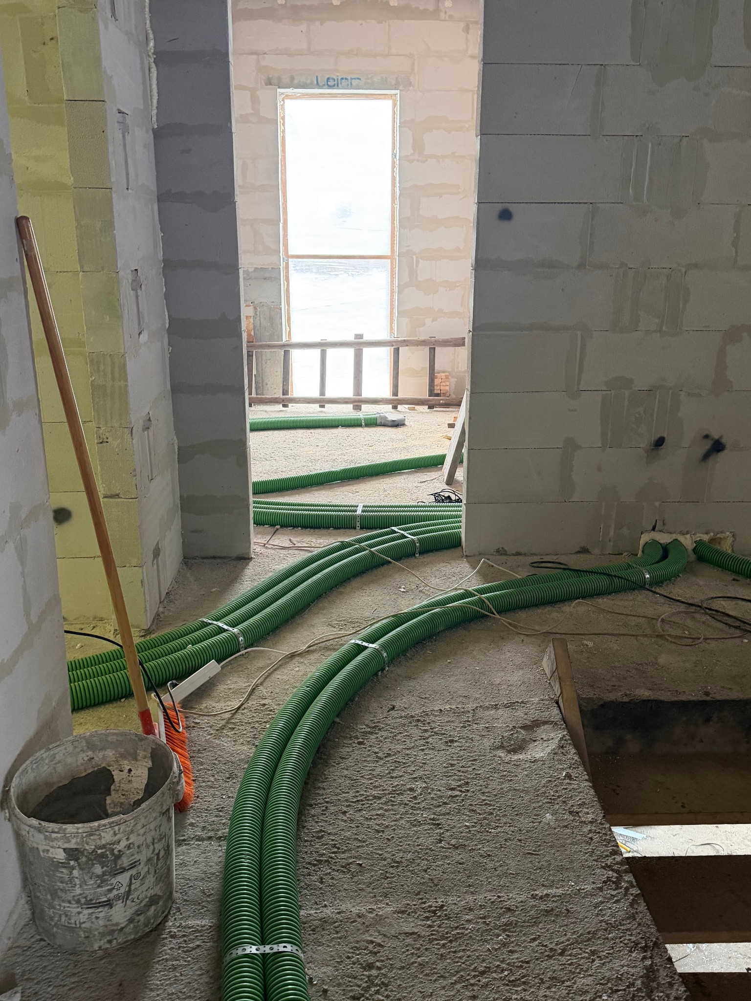 Wnętrze w trakcie budowy z widocznymi zielonymi rurami wentylacyjnymi na podłodze, prowadzącymi w stronę okna. Na pierwszym planie wiadro i miotła.
