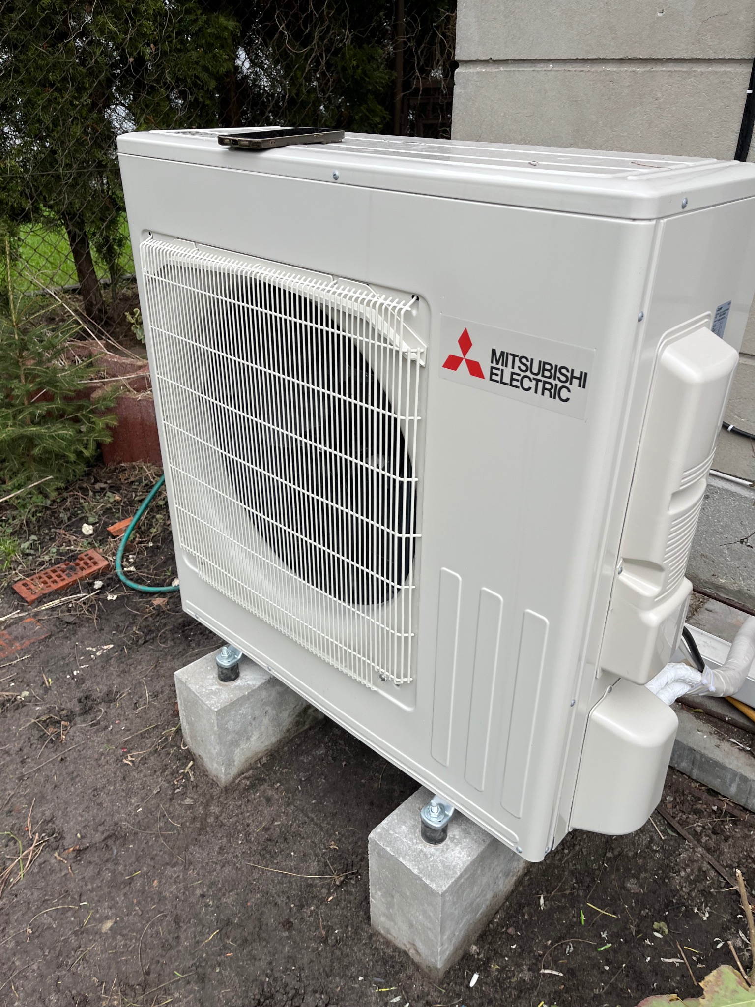 Biała jednostka zewnętrzna klimatyzacji Mitsubishi Electric, umieszczona na betonowych blokach w ogrodzie, z telefonem leżącym na górze. Widoczne elementy instalacji.