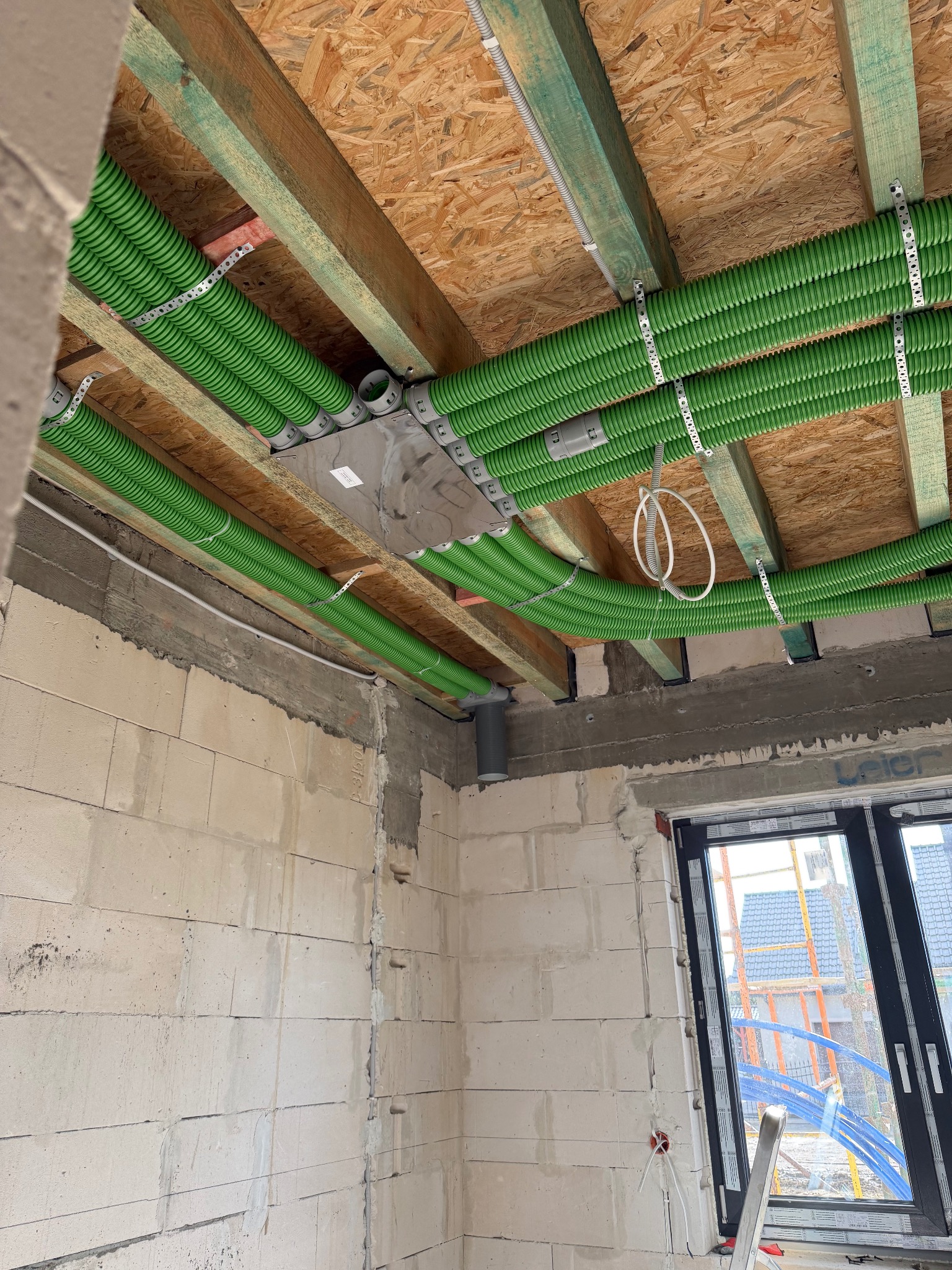 Zielone rury wentylacyjne zamontowane pod drewnianym stropem w surowym wnętrzu budynku z oknem. Widoczne elementy mocujące i skrzynka rozdzielcza.