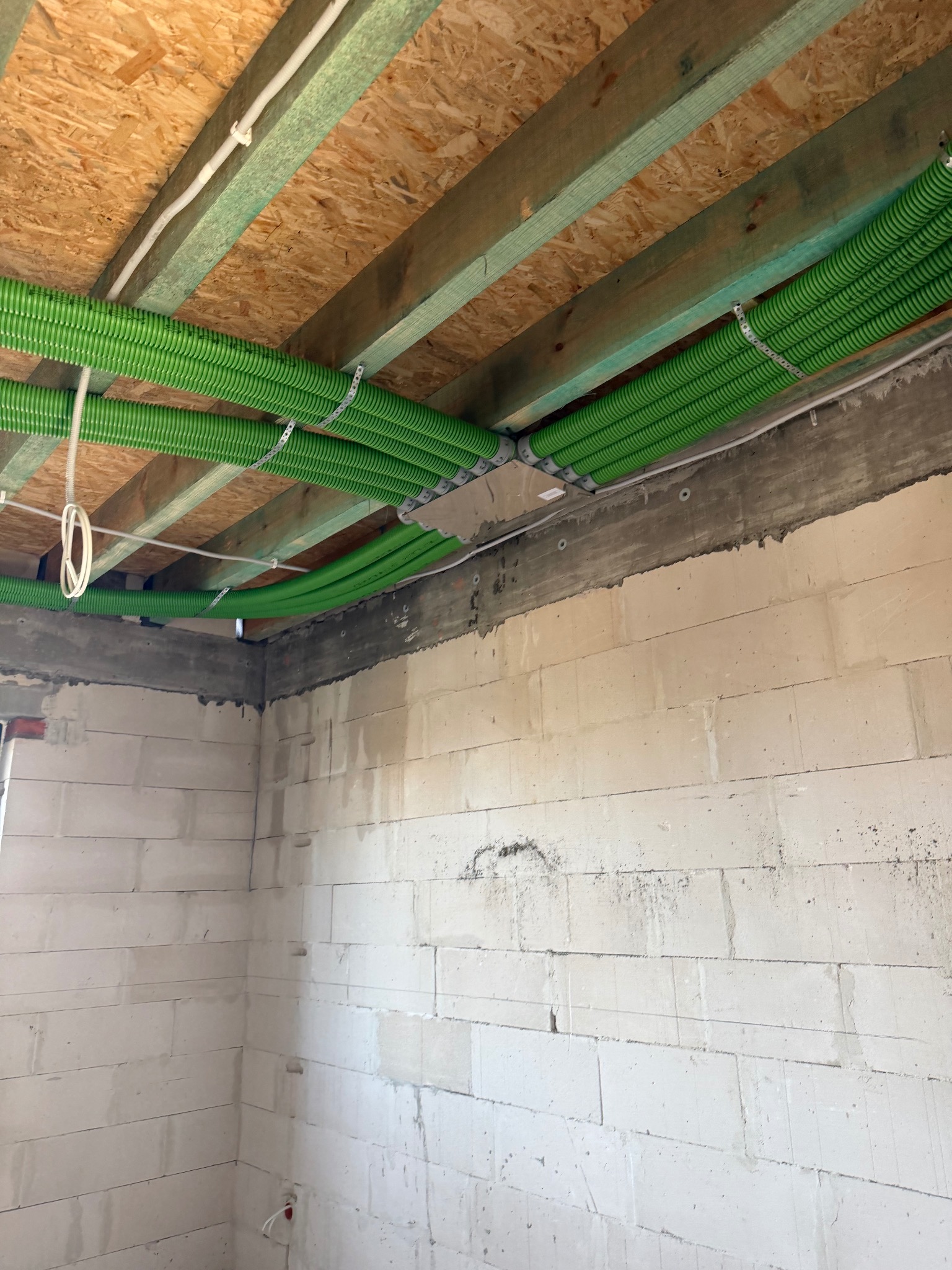 Zielone rury wentylacyjne przymocowane do drewnianego stropu w surowym wnętrzu budynku z białymi ścianami z betonu komórkowego. Widoczne elementy instalacji elektrycznej.