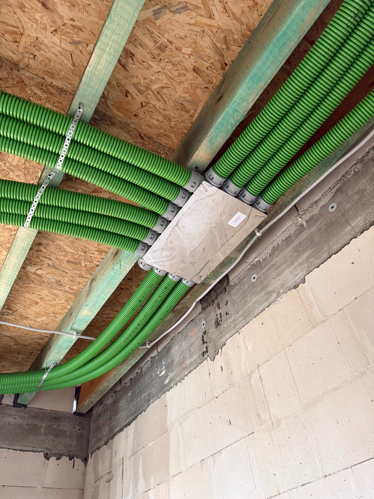 Zielone, karbowane rury wentylacyjne przymocowane do drewnianego stropu z płyt OSB, z widoczną skrzynką rozdzielczą i fragmentem surowej ściany z betonu komórkowego.