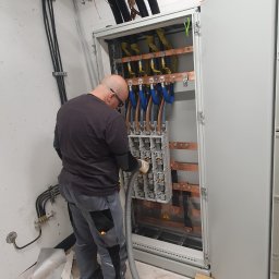 Elektron - Elektryk w okularach czyszczący wnętrze rozdzielnicy elektrycznej za pomocą odkurzacza przemysłowego, widoczne kable i szyny zbiorcze w skrzynce oraz drabinki kablowe pod sufitem.