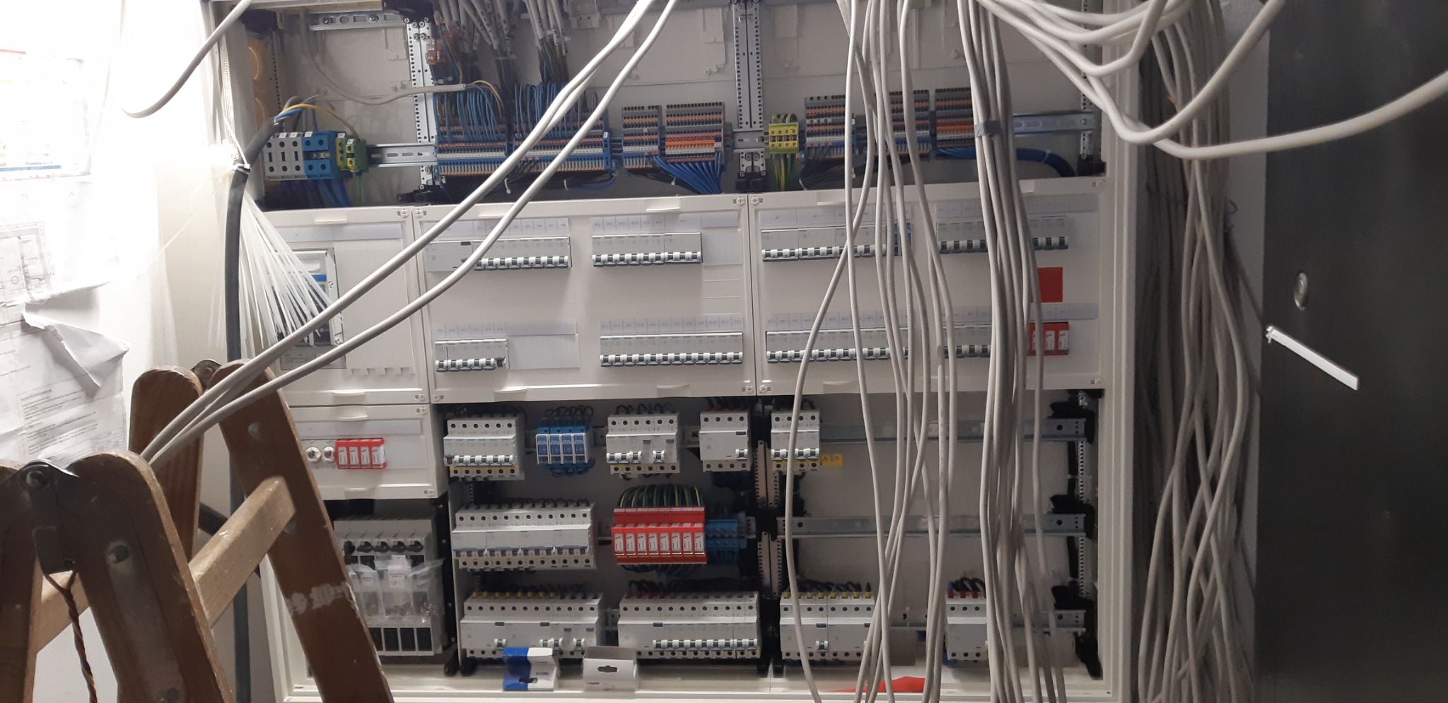 Otwarta szafa rozdzielcza z widocznymi bezpiecznikami, przewodami i listwami zaciskowymi, częściowo zasłonięta przez drewnianą drabinę.