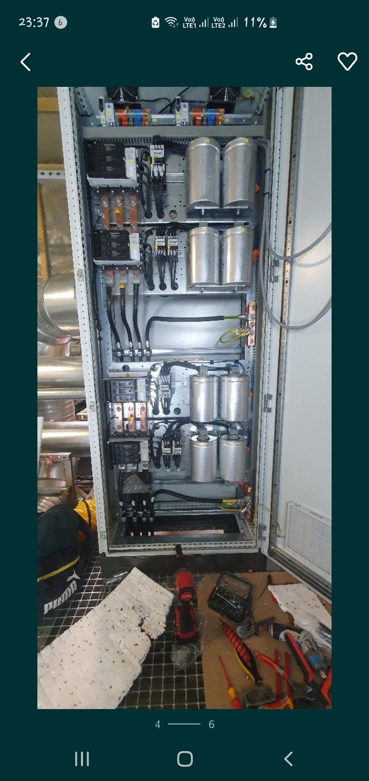 Otwarta szafa elektryczna z widocznymi kondensatorami, zabezpieczeniami i okablowaniem, na podłodze narzędzia i resztki materiałów izolacyjnych.