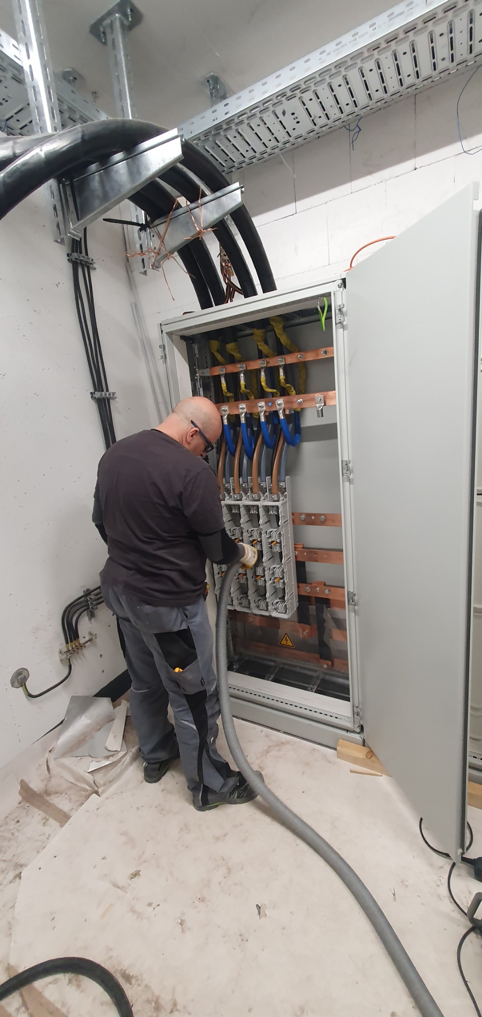 Elektryk w okularach czyszczący wnętrze rozdzielnicy elektrycznej za pomocą odkurzacza przemysłowego, widoczne kable i szyny zbiorcze w skrzynce oraz drabinki kablowe pod sufitem.
