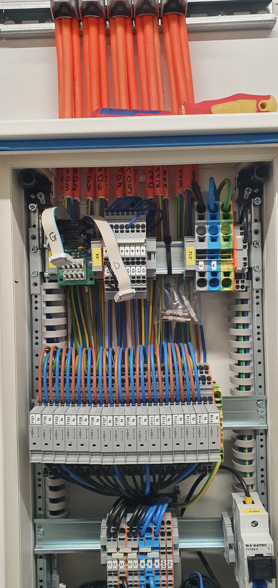 Rozdzielnica elektryczna z uporządkowanymi przewodami, bezpiecznikami i modułami, widoczne oznaczenia przewodów i elementów, narzędzia leżące na górze szafki.