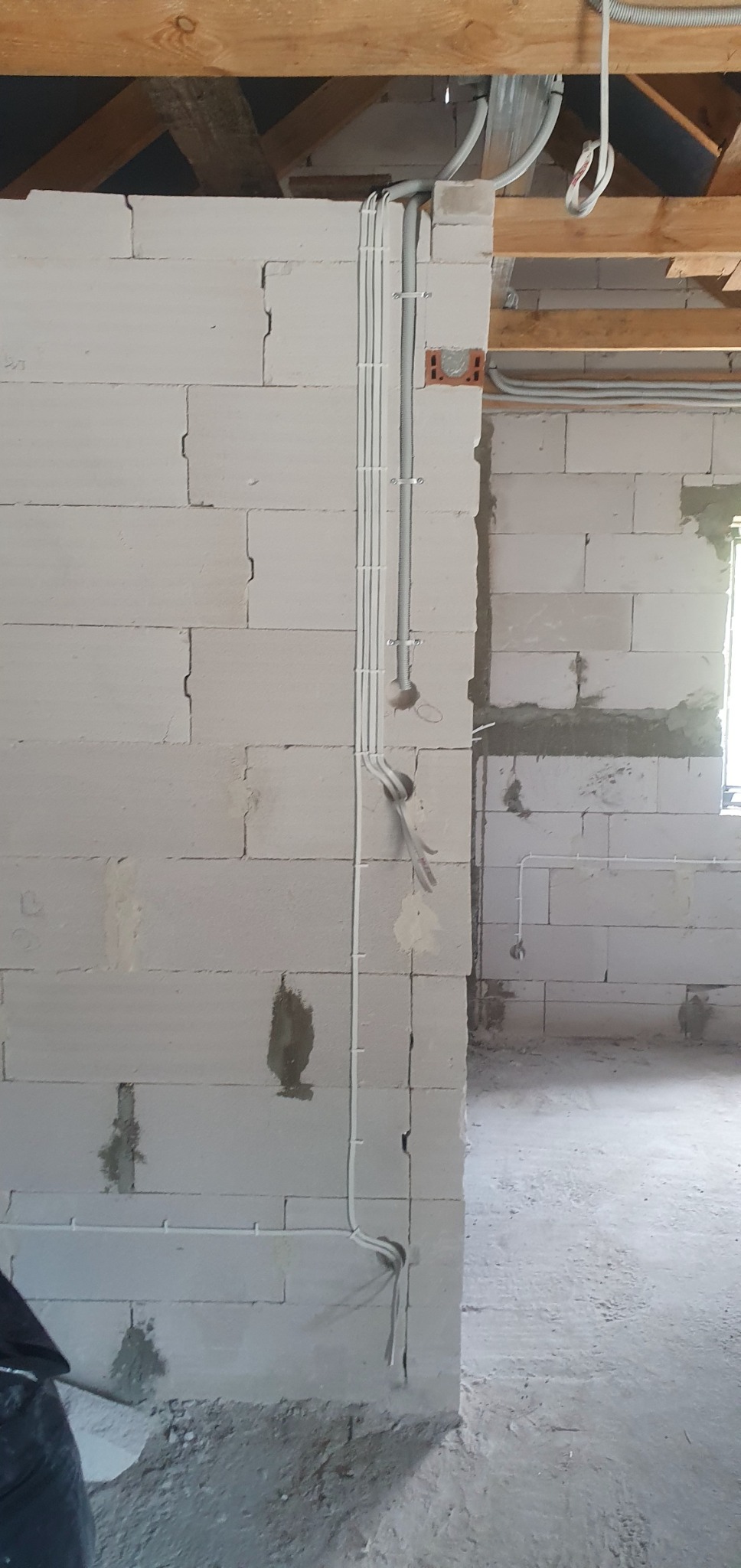 Ściana z betonu komórkowego w trakcie prac elektrycznych: pionowe rury instalacyjne przymocowane klamrami, puszka instalacyjna wmurowana w ścianę, widoczne przewody wystające ze ściany. Surowy stan...