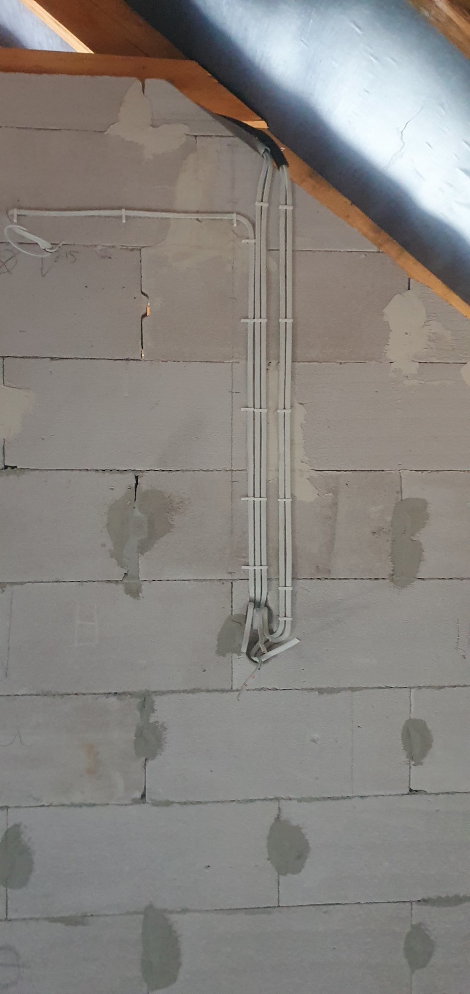 Białe kable elektryczne przymocowane do ściany z bloczków betonowych w surowym stanie, widoczne mocowania i fragment instalacji pod stropem.