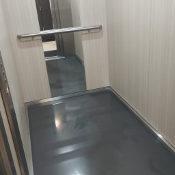 FUNDACJA NOSTRA - Wnętrze windy z szarą podłogą i jasnymi ścianami, widoczny metalowy uchwyt i odbicie drzwi wejściowych.