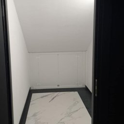 FUNDACJA NOSTRA - Wnętrze małego pomieszczenia z białymi ścianami i podłogą z białego marmuru, widoczne przez otwarte czarne drzwi.