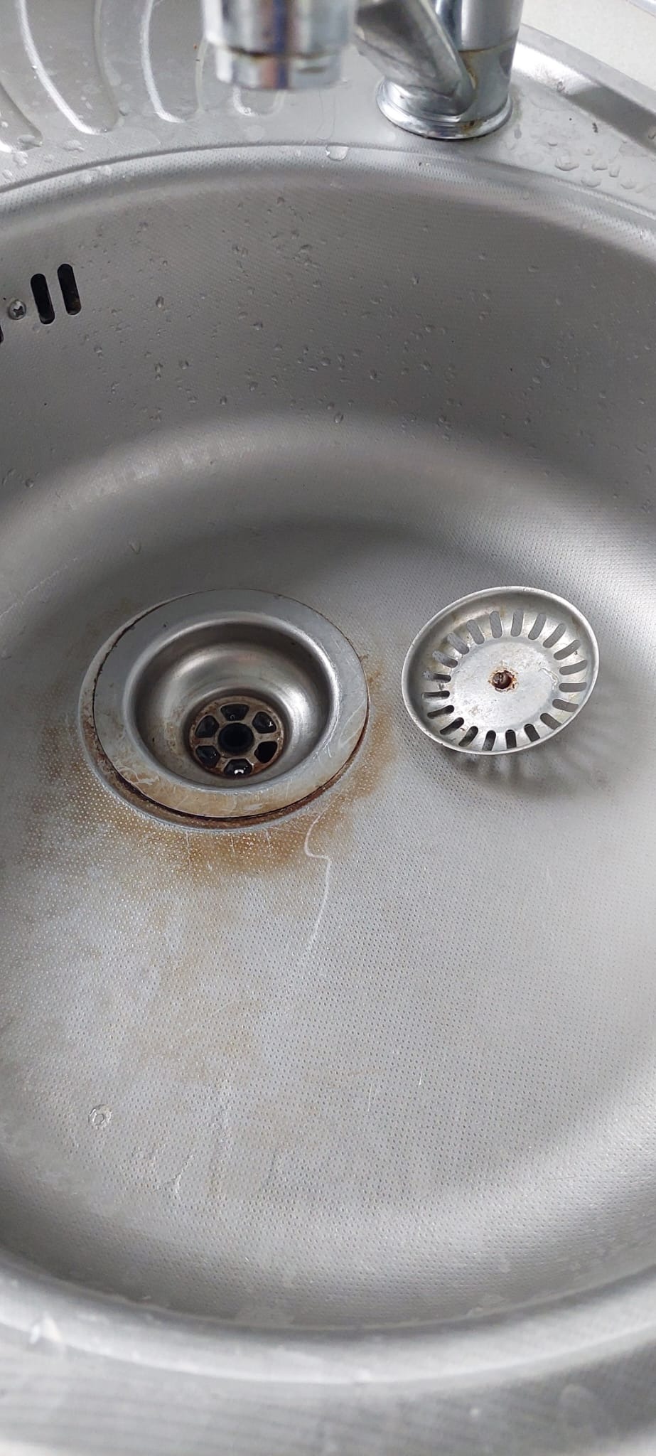Metalowy zlew kuchenny z widocznymi śladami rdzy wokół odpływu i wyjętym sitkiem.