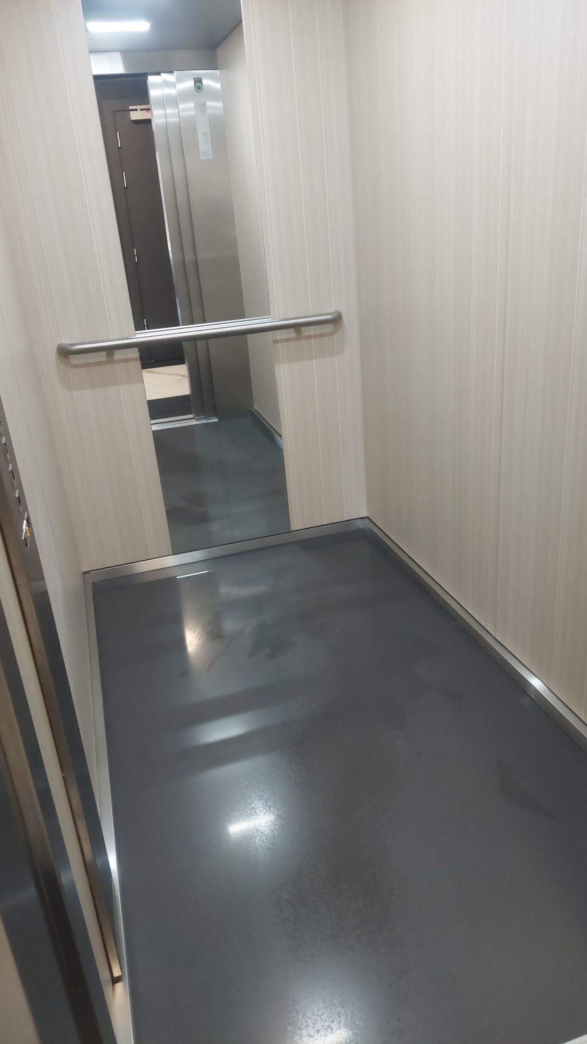 Wnętrze windy z szarą podłogą i jasnymi ścianami, widoczny metalowy uchwyt i odbicie drzwi wejściowych.