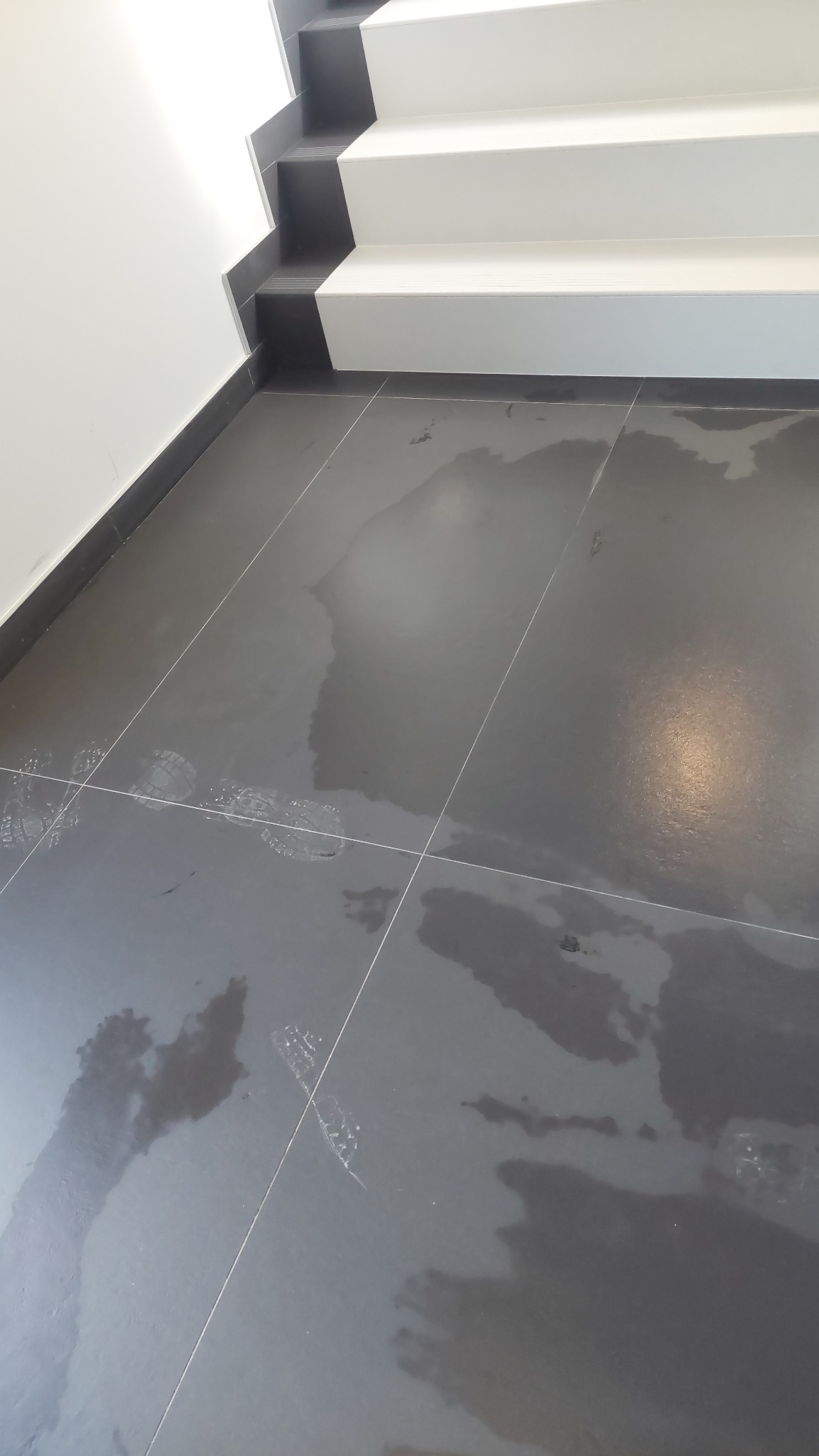 Świeżo umyta, mokra, ciemnoszara podłoga z płytek pod schodami, widoczne ślady stóp.