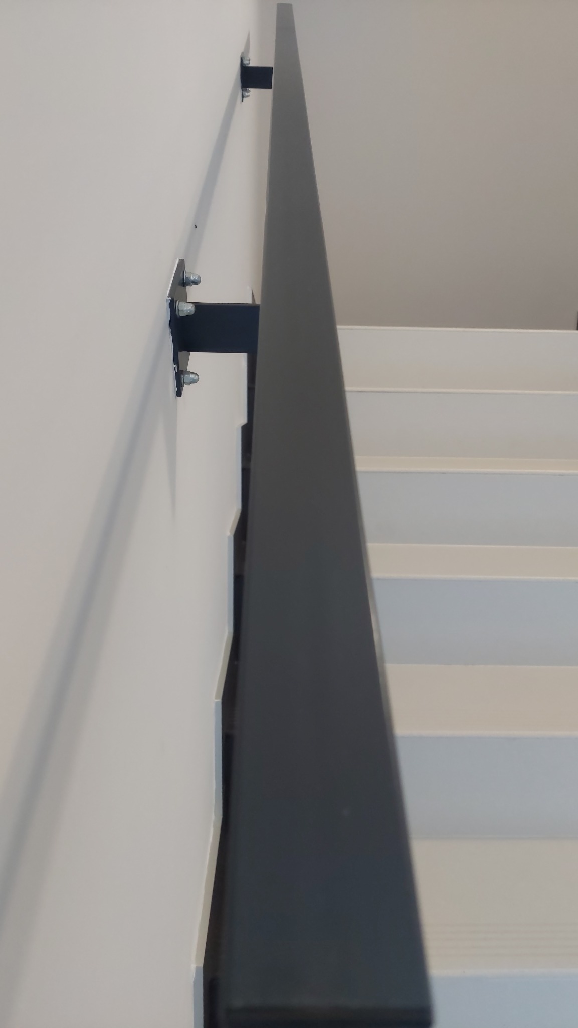 Szczegółowe ujęcie czarnej, metalowej poręczy schodowej przymocowanej do białej ściany za pomocą srebrnych śrub, widoczne białe stopnie schodów.