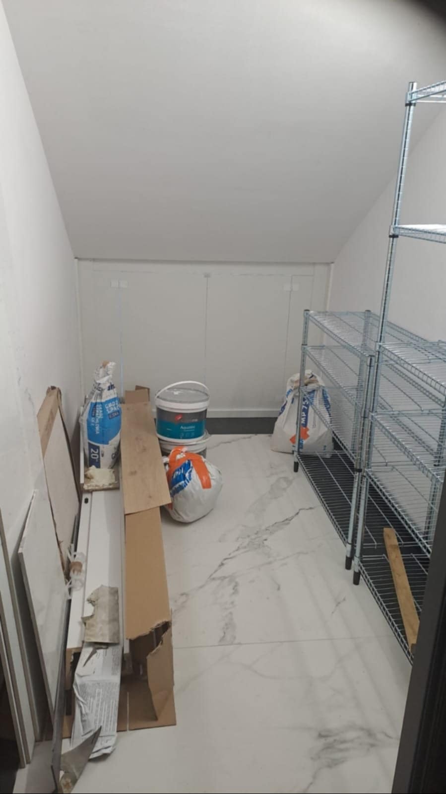 Pomieszczenie gospodarcze z białymi ścianami, marmurową podłogą, metalowymi regałami i materiałami budowlanymi: worki z zaprawą, puszki z farbą i kartony.