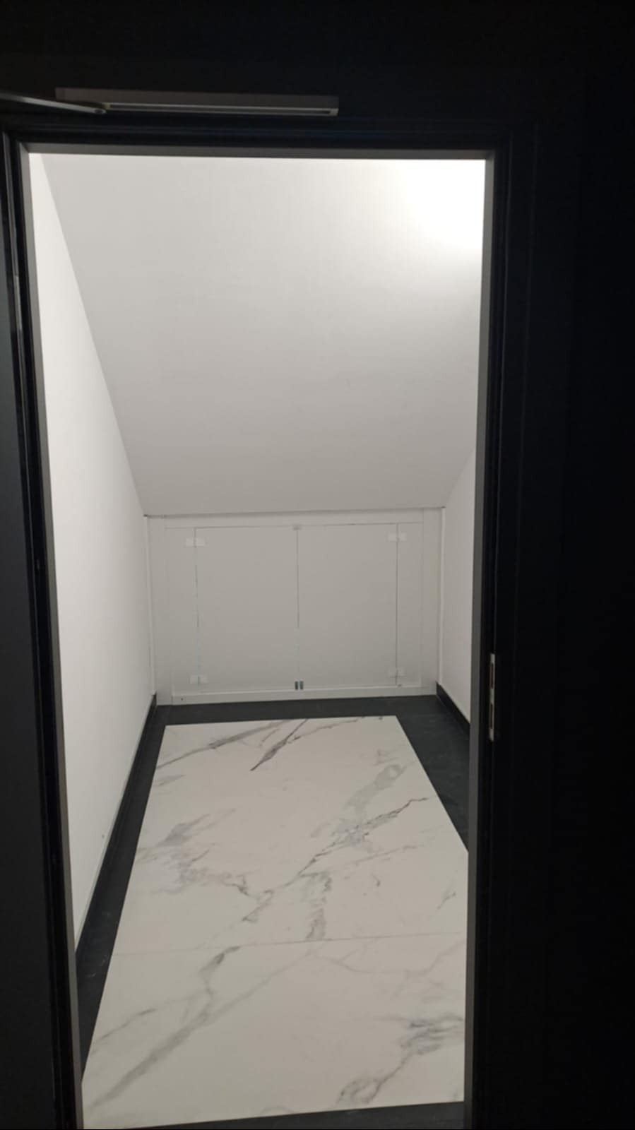 Wnętrze małego pomieszczenia z białymi ścianami i podłogą z białego marmuru, widoczne przez otwarte czarne drzwi.