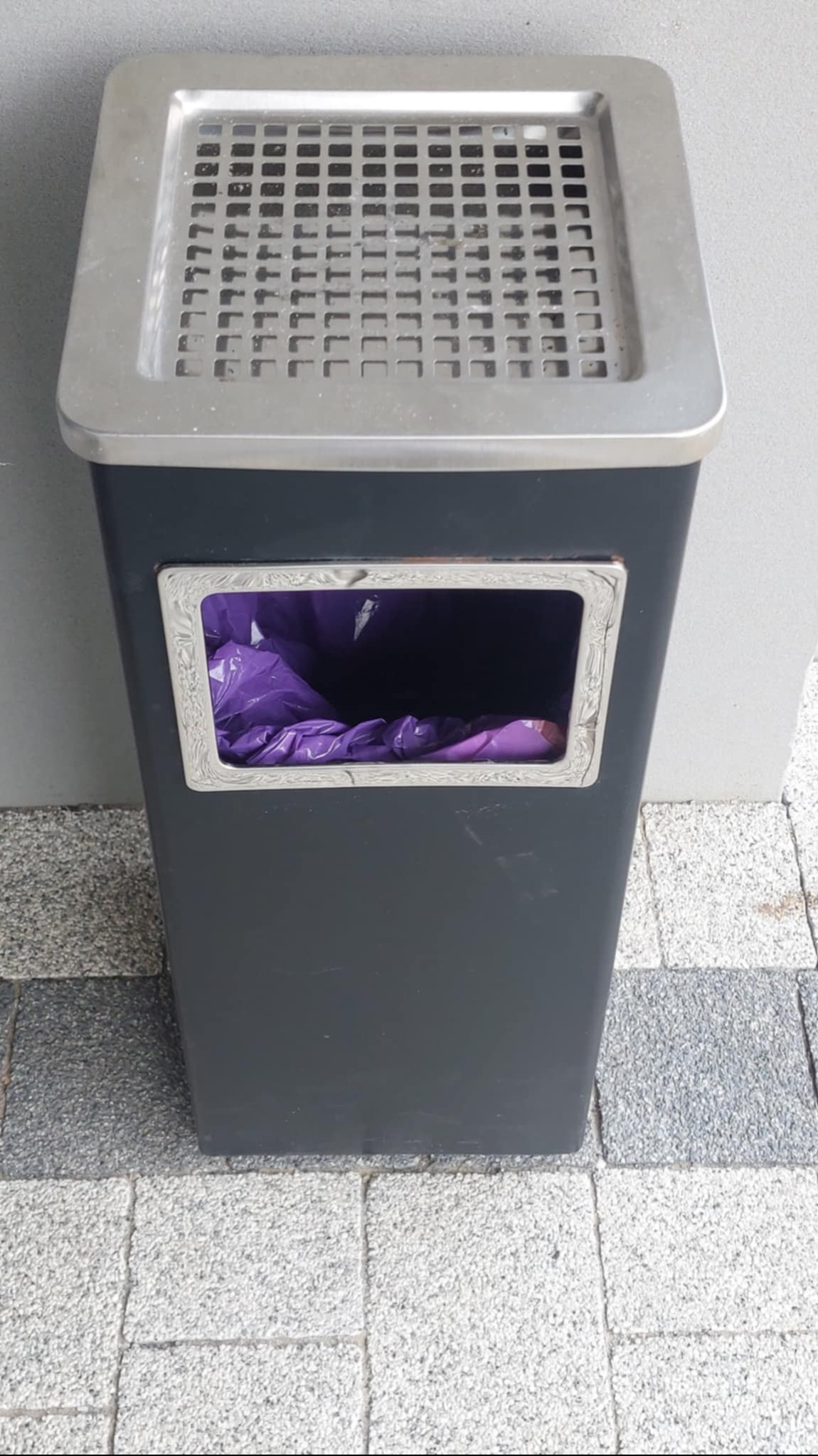 Szary kosz na śmieci z metalową kratką u góry i purpurowym workiem widocznym przez okienko, stojący na szarej kostce brukowej.