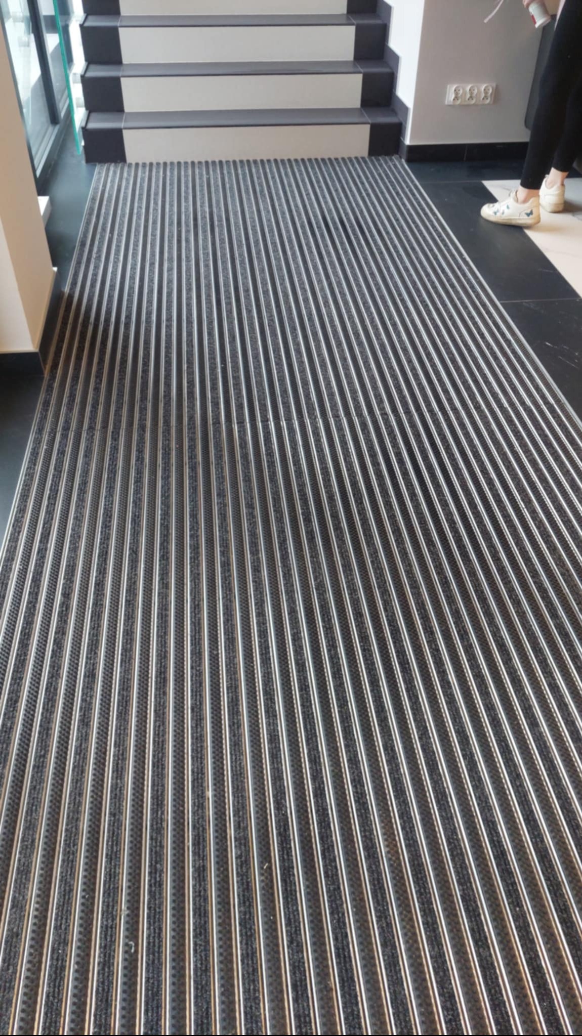 Szara wycieraczka systemowa z aluminiowymi listwami przed schodami wejściowymi do budynku.
