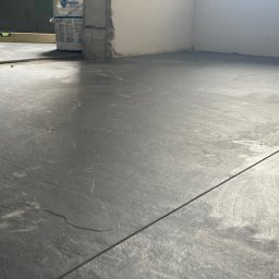 Raman Radzionchyk - Szare płytki podłogowe imitujące kamień, ułożone w pomieszczeniu w trakcie prac wykończeniowych, widoczny poziomica i wiadro z gładzią.