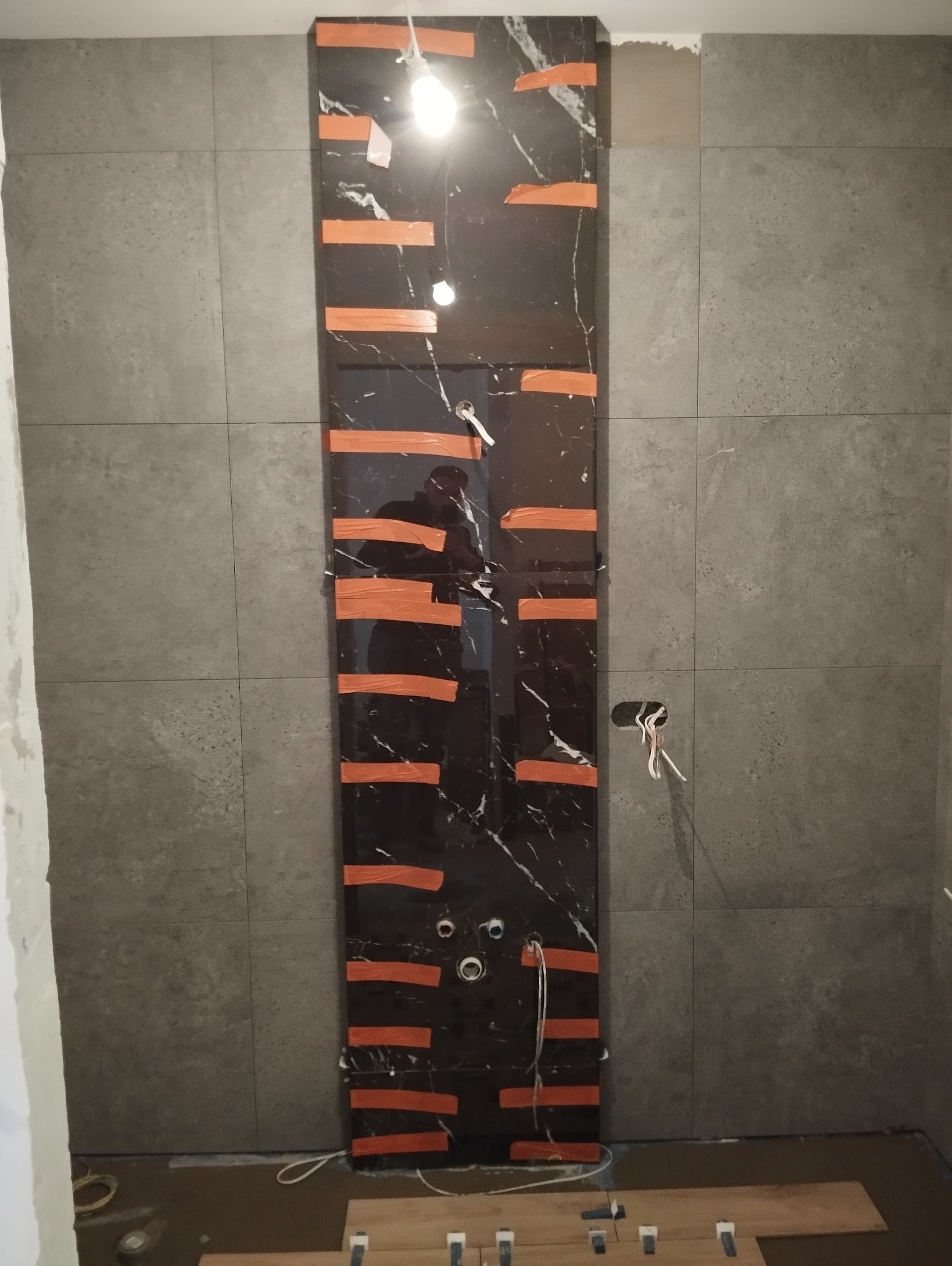 Fragment ściany w łazience z ciemną, marmurkową płytką, zabezpieczoną pomarańczową taśmą malarską, widoczne otwory na instalacje, szare płytki na pozostałej części ściany.