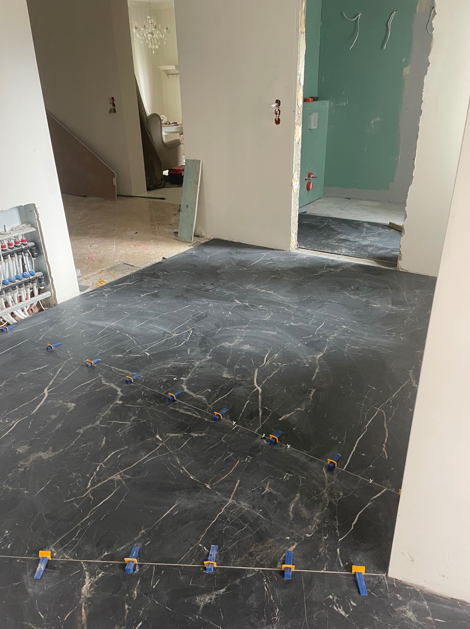 Układanie czarnych płytek podłogowych z białym wzorem marmuru, z użyciem niebieskich i pomarańczowych krzyżyków dystansowych, widoczna instalacja grzewcza podłogowa w tle.