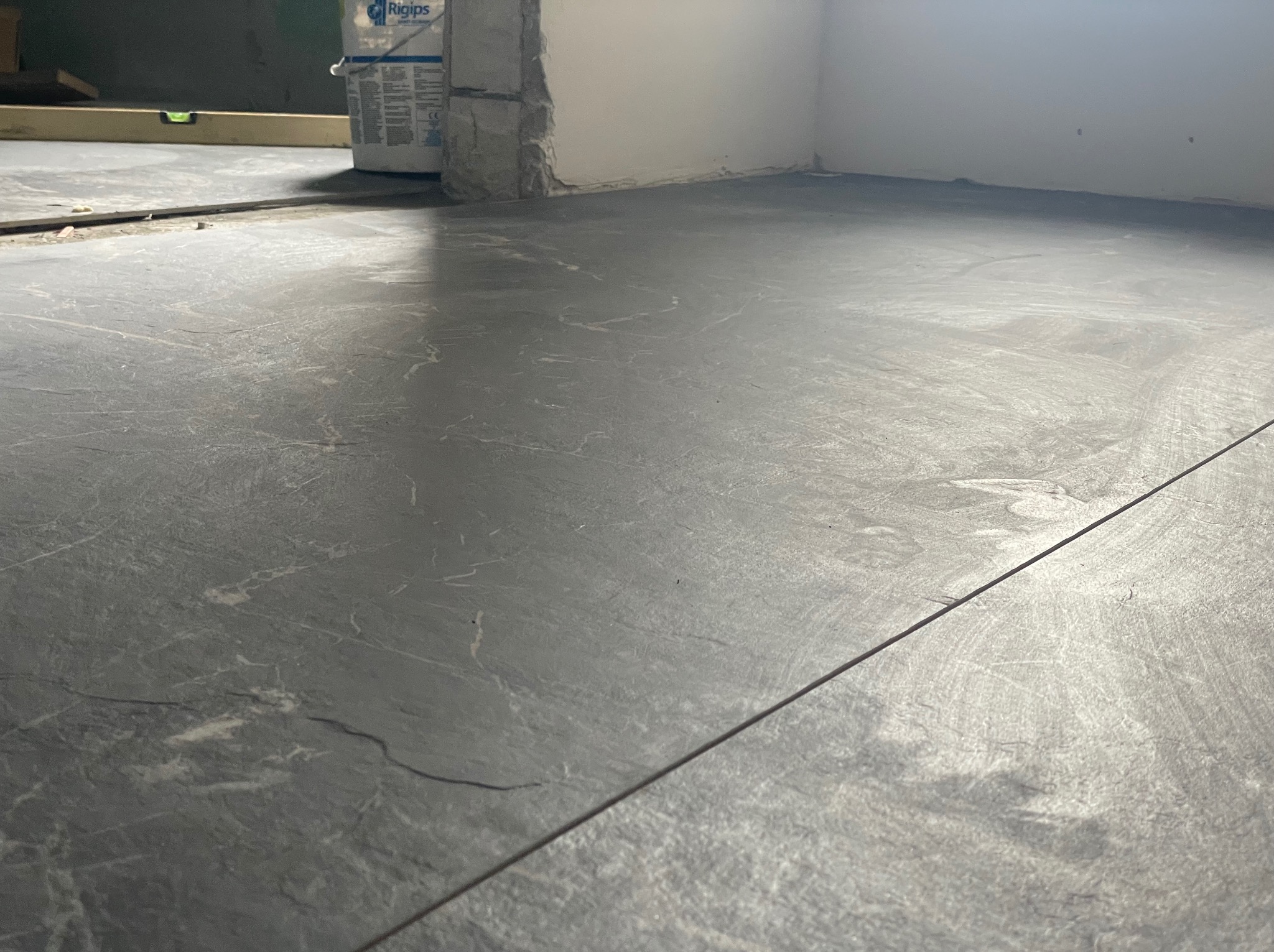Szare płytki podłogowe imitujące kamień, ułożone w pomieszczeniu w trakcie prac wykończeniowych, widoczny poziomica i wiadro z gładzią.