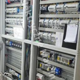 Rozbudowana szafa sterownicza w trakcie montażu, widoczne liczne kable, bezpieczniki i dokumentacja techniczna na czarnej podkładce.