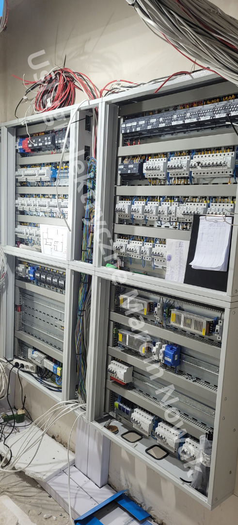 Rozbudowana szafa sterownicza w trakcie montażu, widoczne liczne kable, bezpieczniki i dokumentacja techniczna na czarnej podkładce.