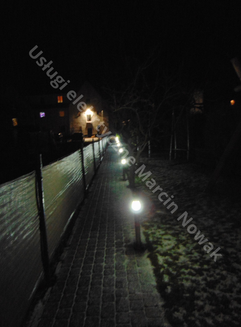 Oświetlona ścieżka z kostki brukowej wzdłuż ogrodzenia, nocne ujęcie z lampami ogrodowymi wzdłuż drogi, widoczny zarys domu w tle.