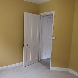 MZ USŁUGI MALARSKIE - Pomieszczenie po malowaniu ścian na kolor musztardowy, widoczne białe drzwi z czarną klamką i listwy przypodłogowe, na podłodze jasna wykładzina.
