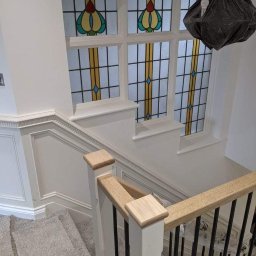 MZ USŁUGI MALARSKIE - Klatka schodowa z drewnianą balustradą, szarym dywanem na schodach i oknem witrażowym z geometrycznym wzorem. Lampa tymczasowo osłonięta czarnym workiem.