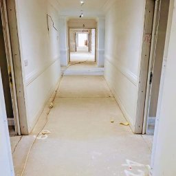 MZ USŁUGI MALARSKIE - Długi korytarz w trakcie remontu, ściany po szpachlowaniu, widoczne ślady po pracach na podłodze, kable elektryczne i białe napisy na posadzce.