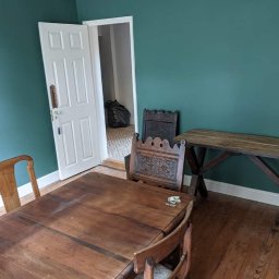 MZ USŁUGI MALARSKIE - Pomieszczenie z zielonymi ścianami po malowaniu, widoczne meble w stylu rustykalnym i otwarte drzwi do sąsiedniego pokoju z wzorzystą podłogą.