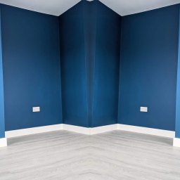 MZ USŁUGI MALARSKIE - Pomalowany na granatowo róg pokoju z białymi listwami przypodłogowymi i panelami podłogowymi imitującymi drewno, widoczne gniazdka elektryczne.