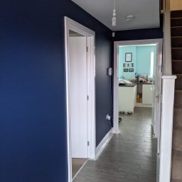 MZ USŁUGI MALARSKIE - Korytarz po remoncie: ściany pomalowane na granatowo, białe listwy przypodłogowe, szare panele podłogowe imitujące drewno, widok na jasną kuchnię w tle.