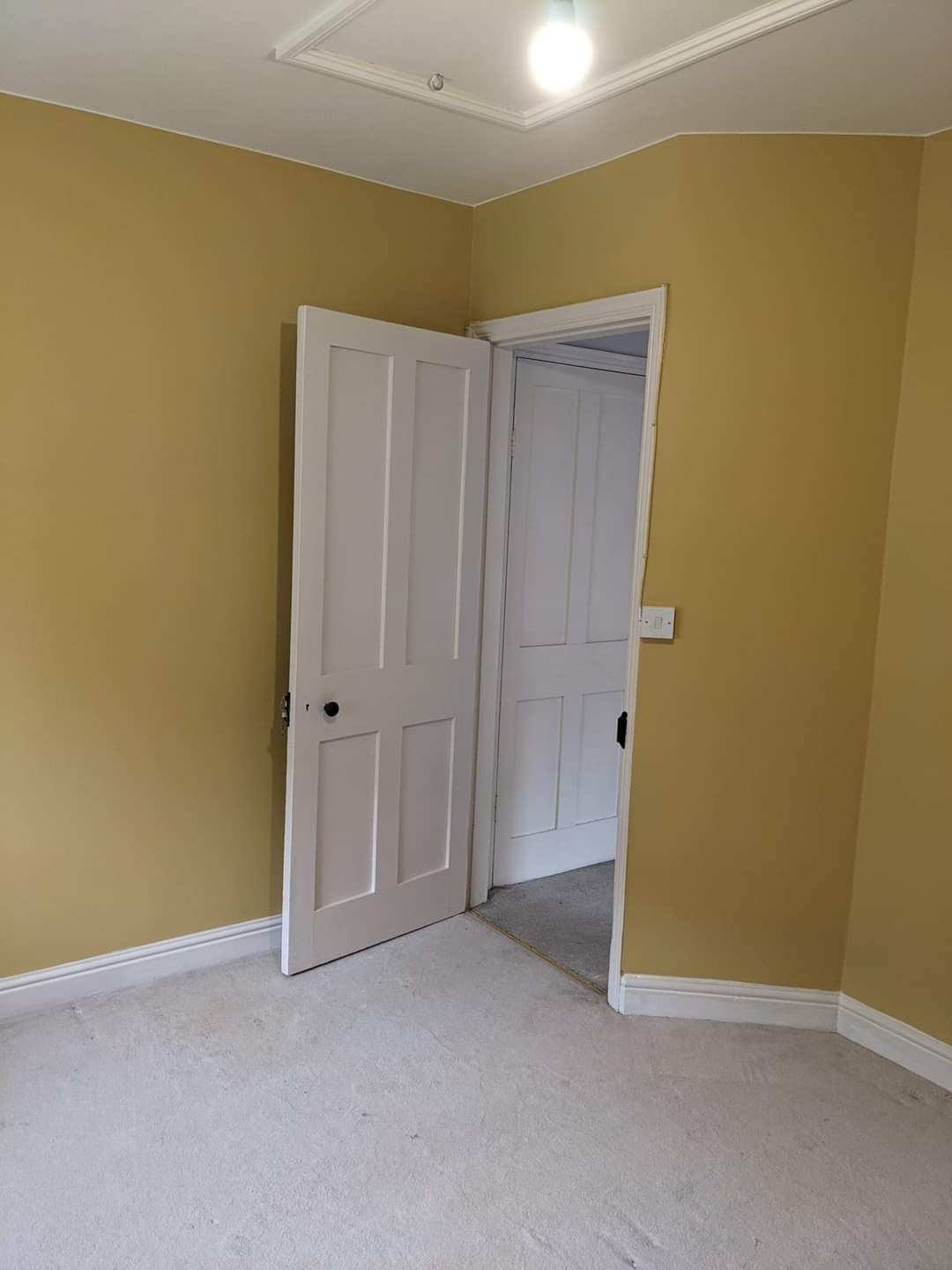 Pomieszczenie po malowaniu ścian na kolor musztardowy, widoczne białe drzwi z czarną klamką i listwy przypodłogowe, na podłodze jasna wykładzina.