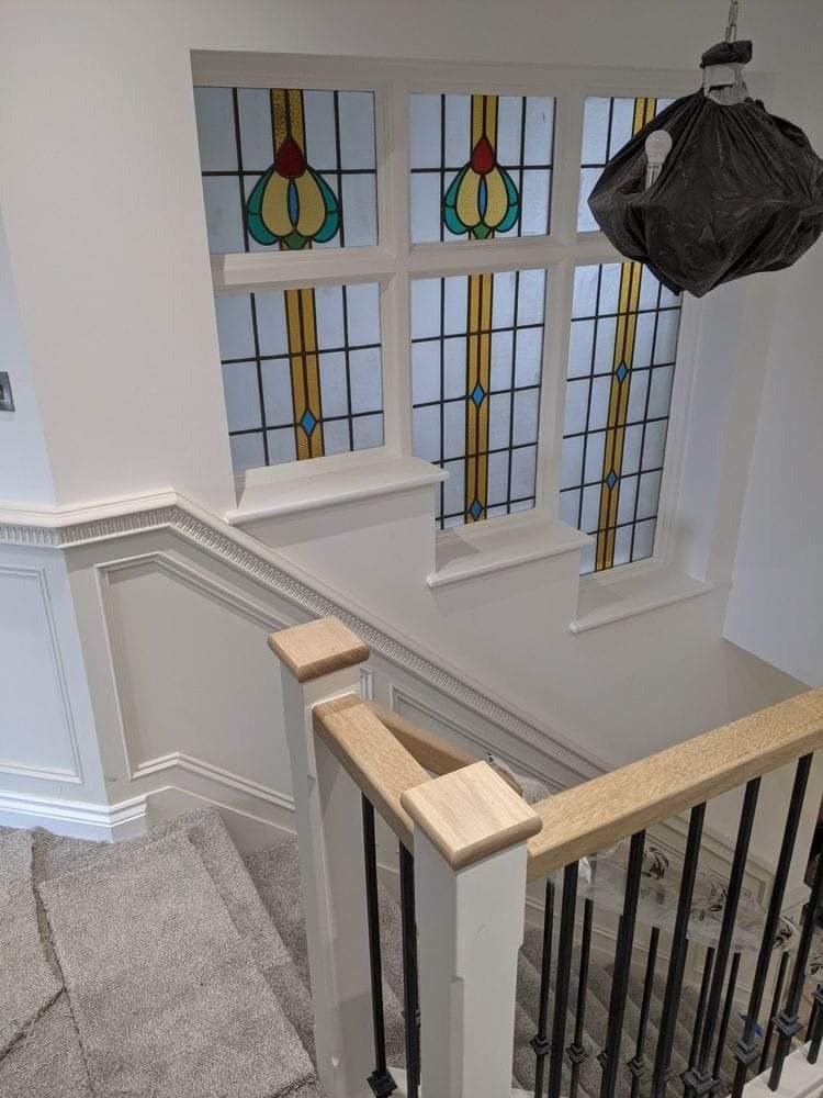 Klatka schodowa z drewnianą balustradą, szarym dywanem na schodach i oknem witrażowym z geometrycznym wzorem. Lampa tymczasowo osłonięta czarnym workiem.