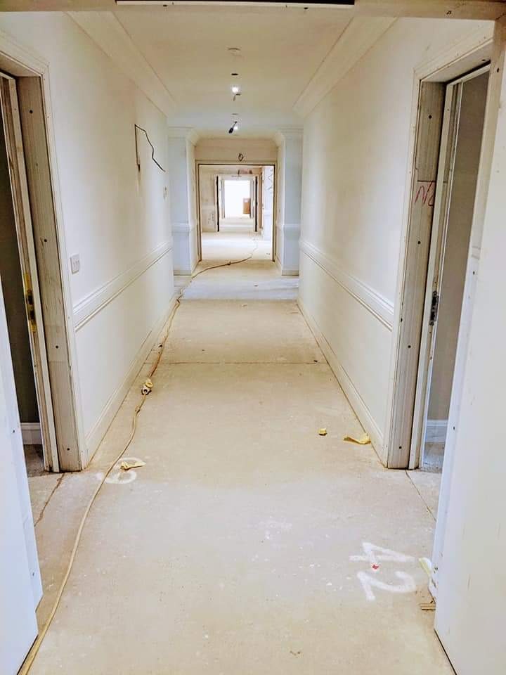 Długi korytarz w trakcie remontu, ściany po szpachlowaniu, widoczne ślady po pracach na podłodze, kable elektryczne i białe napisy na posadzce.