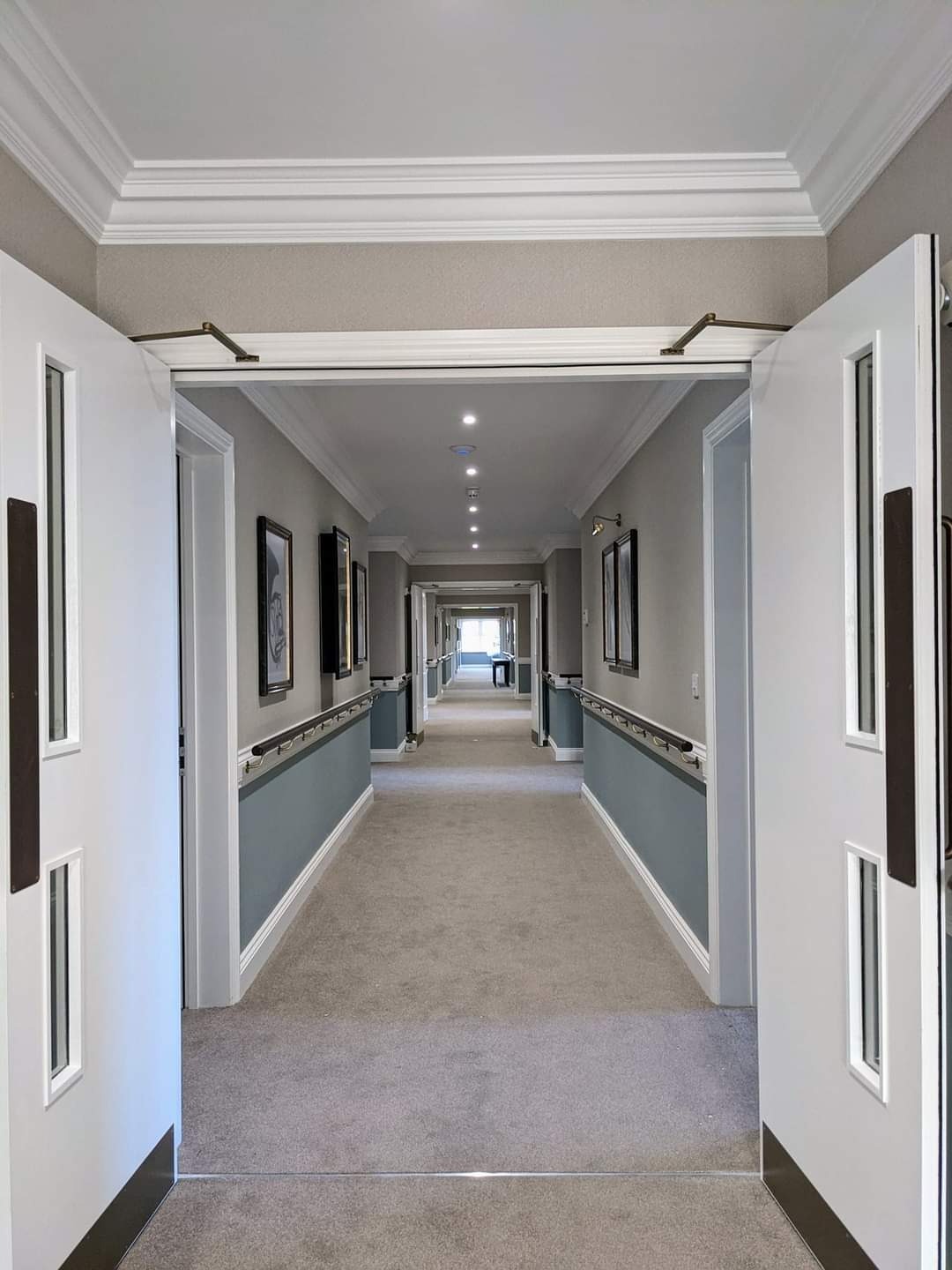 Długi korytarz w budynku użyteczności publicznej z szarą wykładziną, pomalowanymi na szaro ścianami z niebieskimi panelami dolnymi, oświetleniem sufitowym i białymi drzwiami z ciemnymi uchwytami...