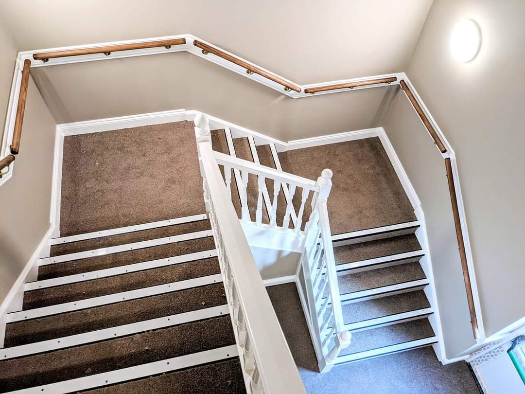 Widok z góry na klatkę schodową z brązową wykładziną i białymi listwami oraz drewnianą poręczą, świeżo po remoncie, z widocznym oświetleniem sufitowym.