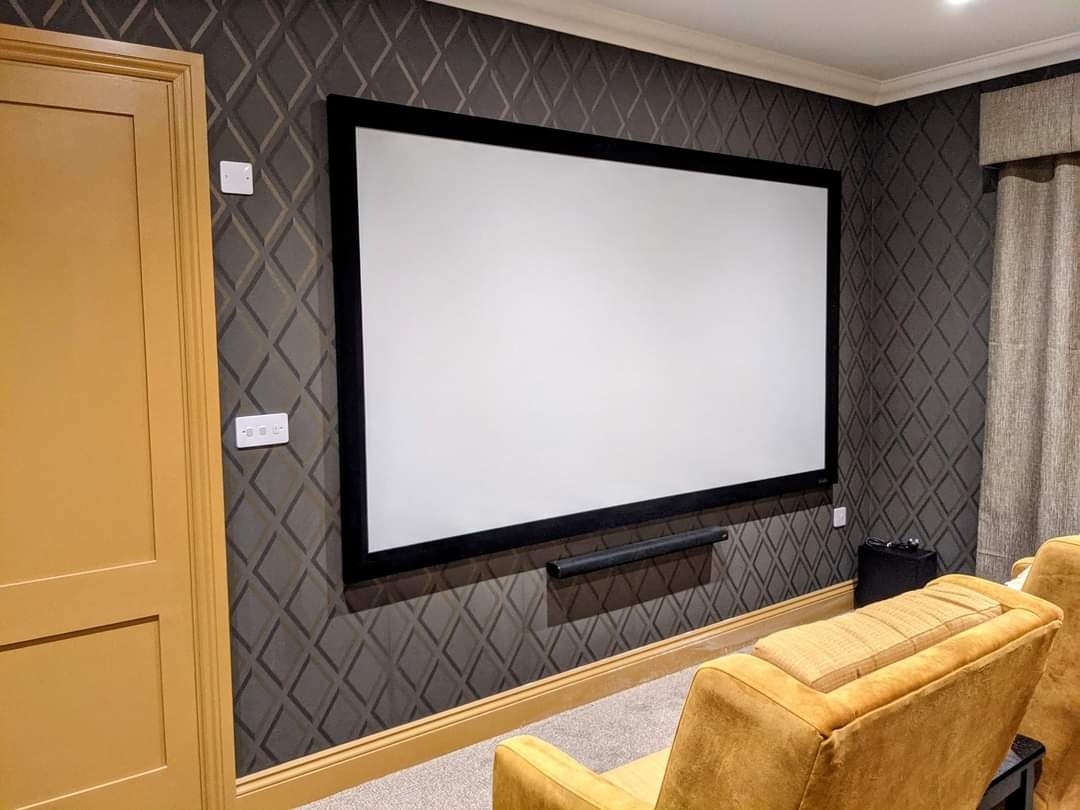 Domowe kino z dużym ekranem projekcyjnym na ścianie z geometrycznym wzorem, widoczne krzesła w kolorze musztardowym, listwa głośnikowa pod ekranem.