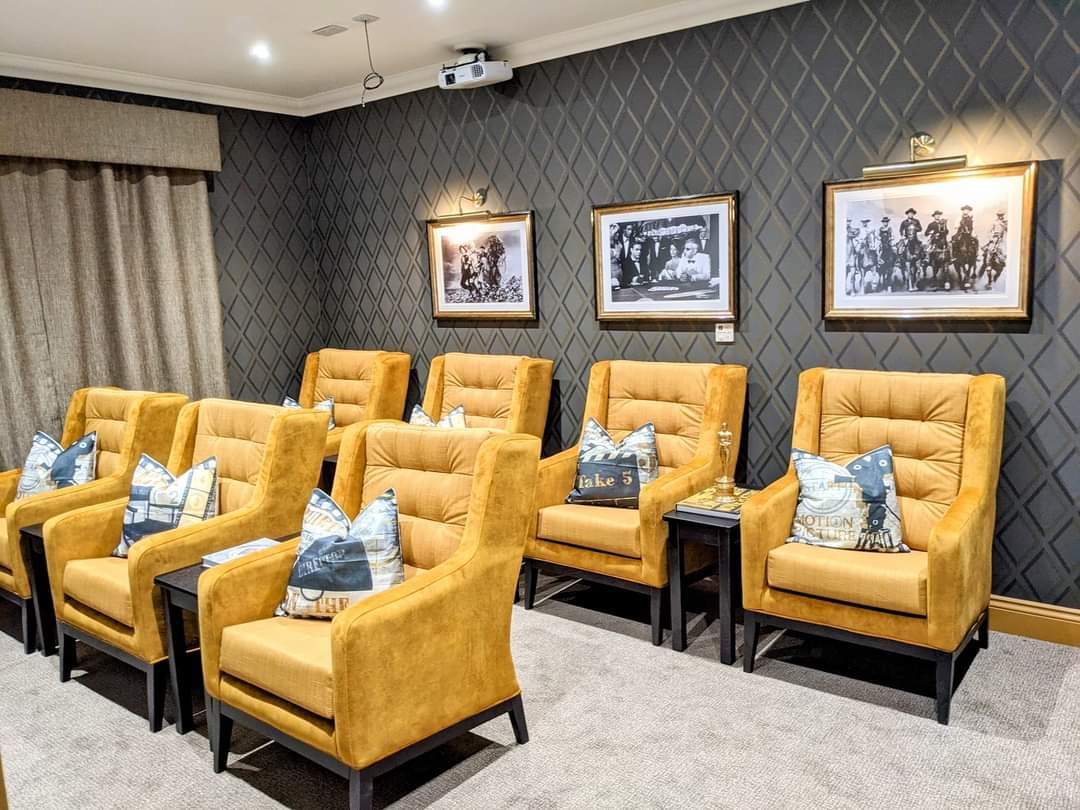 Sala kinowa z rzędem musztardowych foteli z poduszkami filmowymi, czarne stoliki boczne, ściana w romby, zdjęcia w złotych ramach i projektor sufitowy.