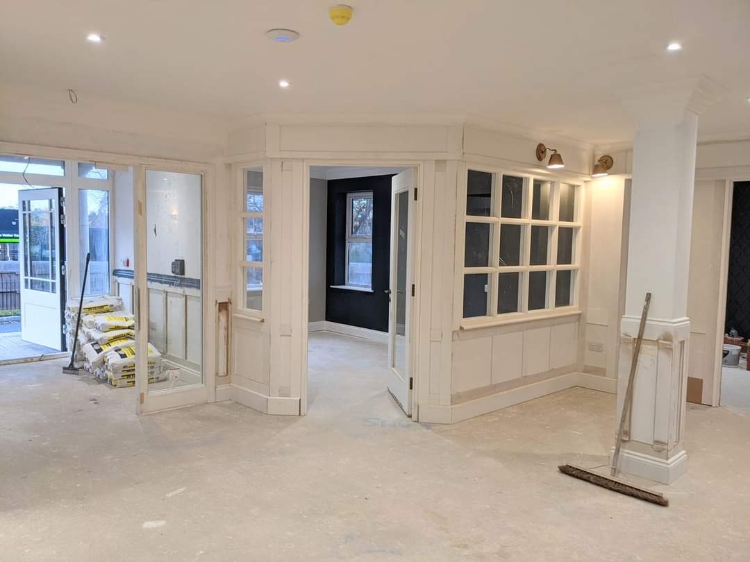 Wnętrze w trakcie prac wykończeniowych: białe ściany z drewnianą boazerią, worki z zaprawą przy drzwiach, surowa podłoga i widoczne ślady szpachlowania.