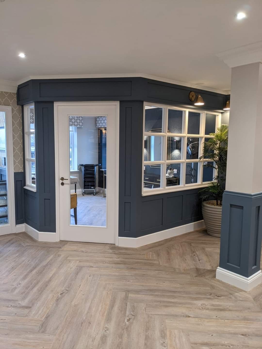 Wykończone wnętrze z ciemnogranatową ścianą z panelami, białymi ramami okiennymi i szklanymi drzwiami, parkiet ułożony w jodełkę, donica z rośliną w rogu.