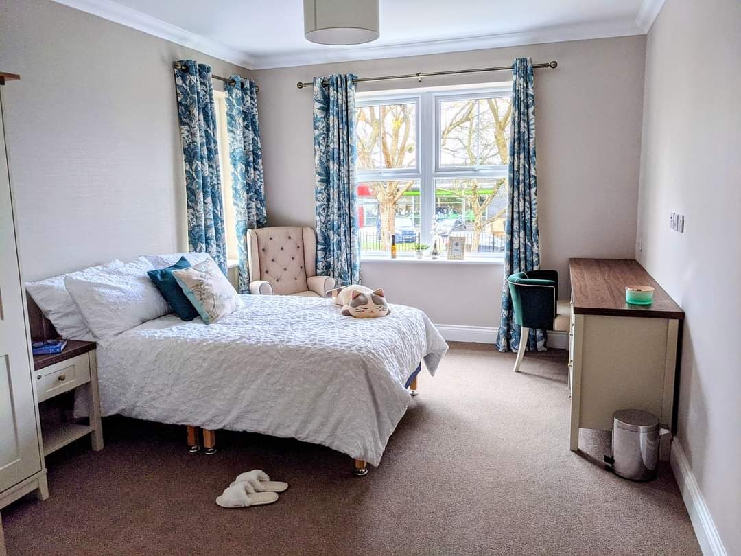 Jasne wnętrze sypialni z łóżkiem, biurkiem i fotelami, ściany pomalowane na jasny odcień szarości, dekoracyjne zasłony w kwiatowy wzór.