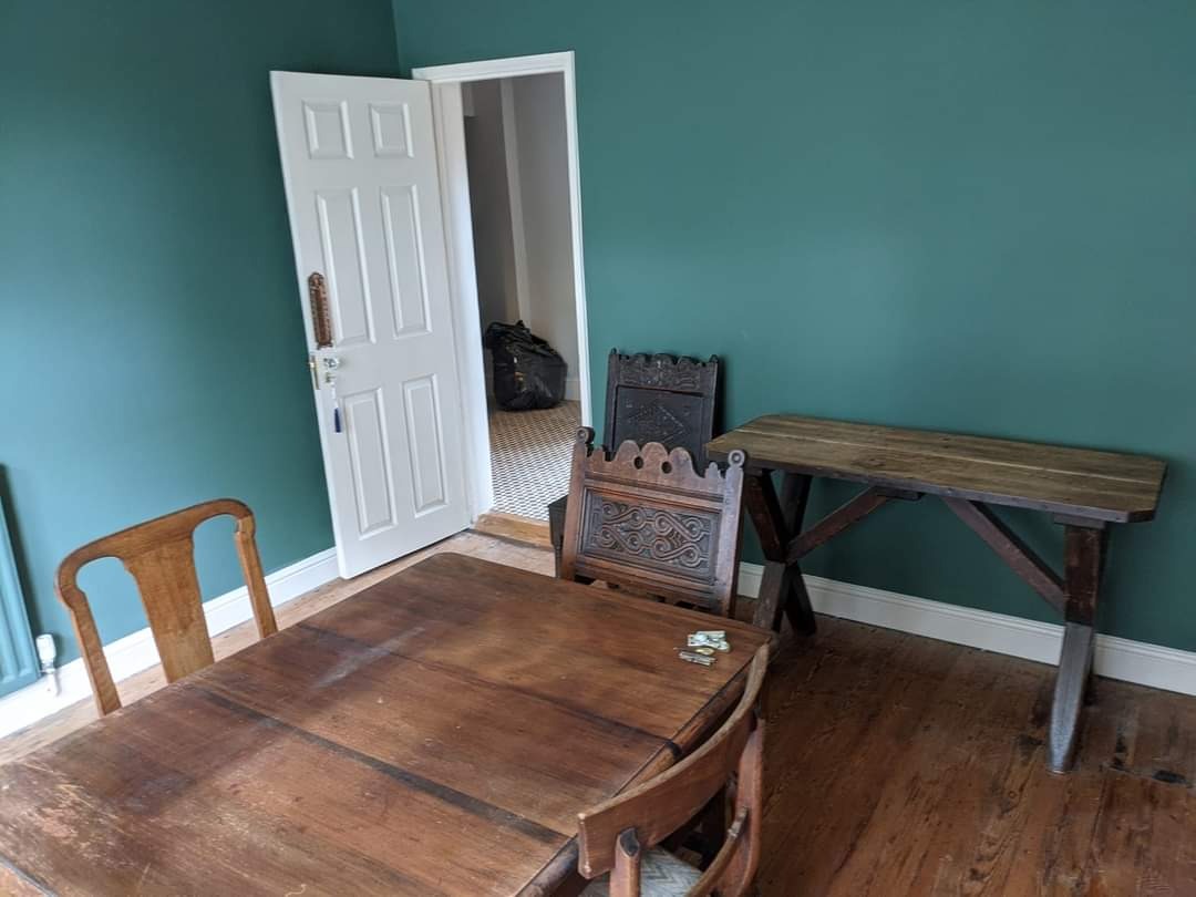 Pomieszczenie z zielonymi ścianami po malowaniu, widoczne meble w stylu rustykalnym i otwarte drzwi do sąsiedniego pokoju z wzorzystą podłogą.